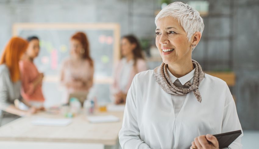 A woman in a white lab coat holds a tablet while other woman can be seen in the background.